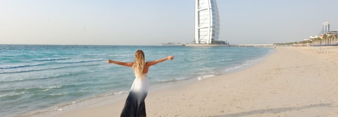 A carefree woman in a flowing dress strolls along the Dubai beach with Burj Al Arab in view.